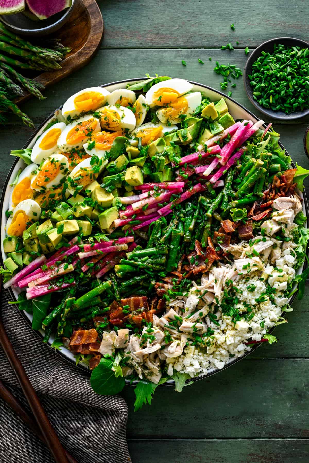 Overhead view of spring cobb salad with hard boiled egg, avocado, radish, asparagus, bacon, chicken and feta cheese in rows.