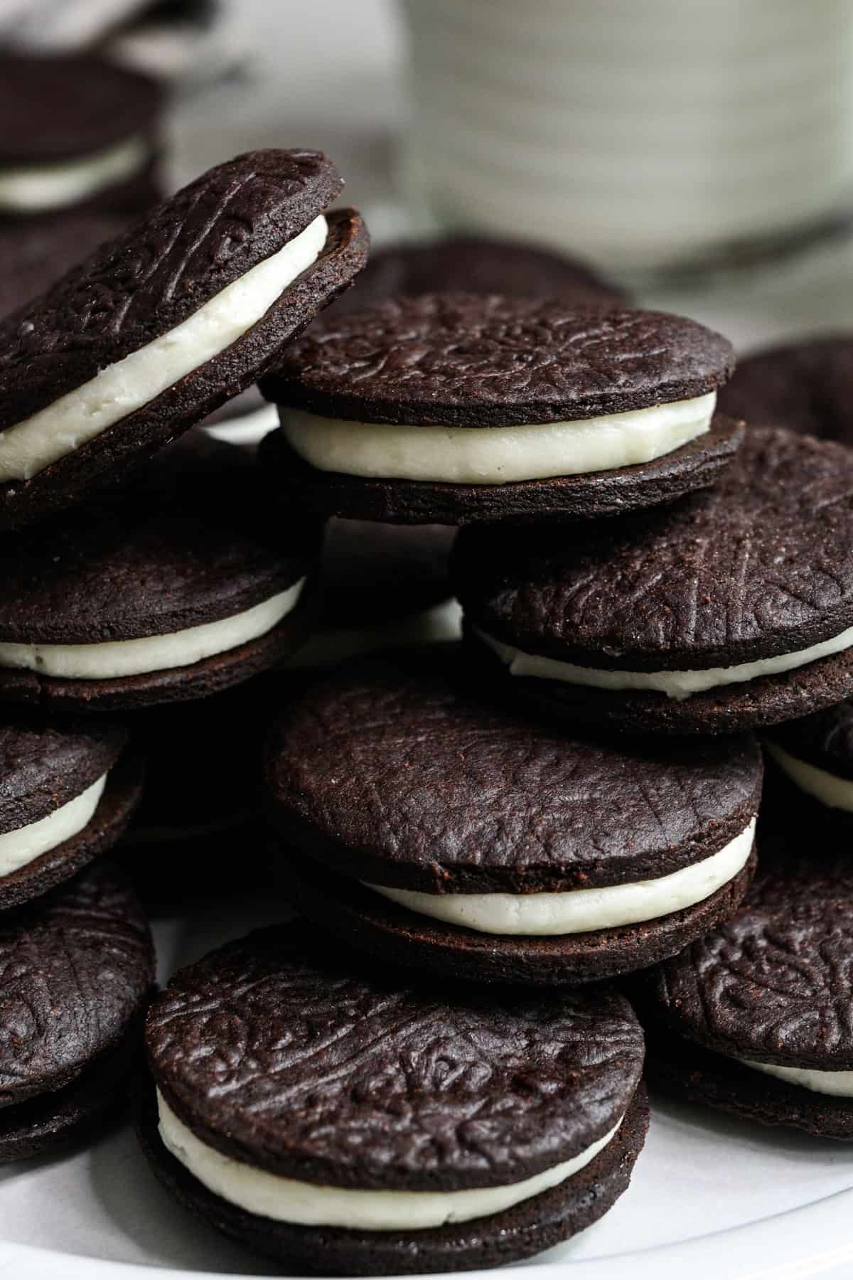 Close up side view of homemade oreos stacked on a white plate with a glass of milk behind.