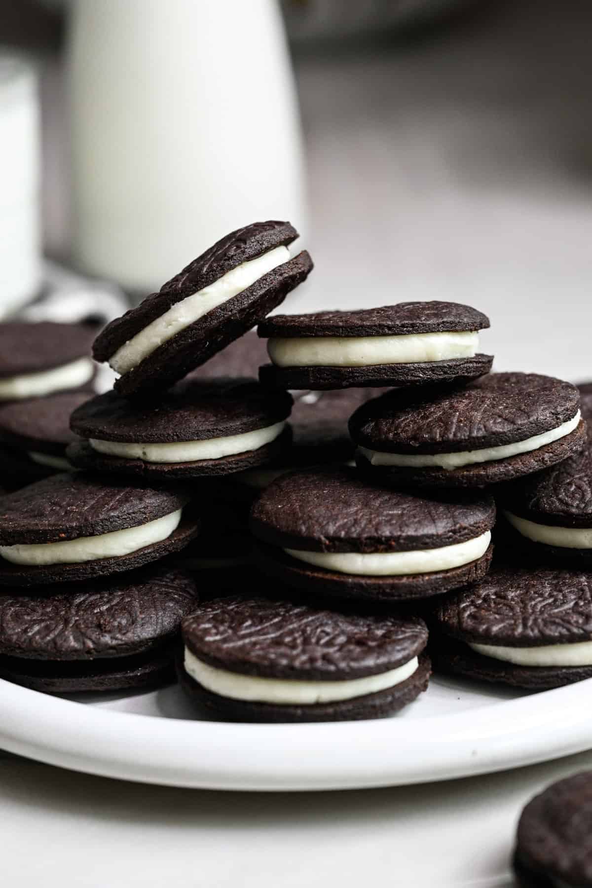 Close up side view of homemade oreos stacked on a white plate with a glass of milk behind.