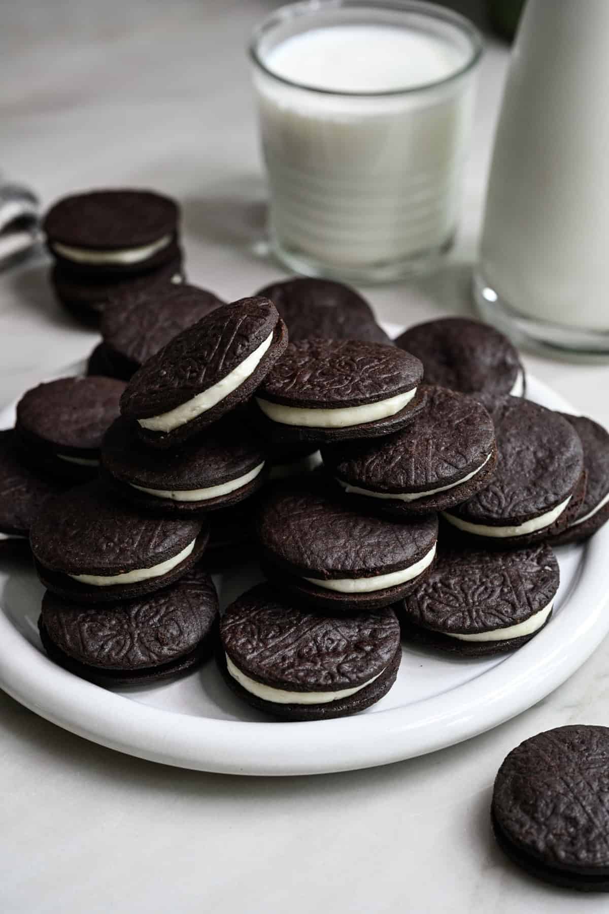 Close up side view of homemade oreos stacked on a white plate with a glass of milk behind.