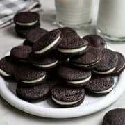 Close up side view of homemade oreos stacked on a white plate with a glass of milk behind.