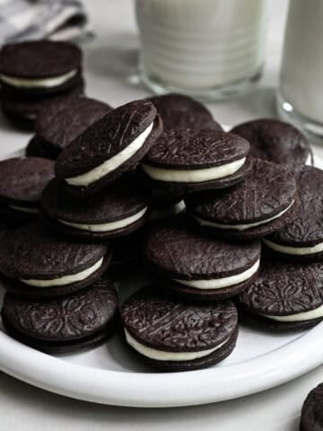 Close up side view of homemade oreos stacked on a white plate with a glass of milk behind.