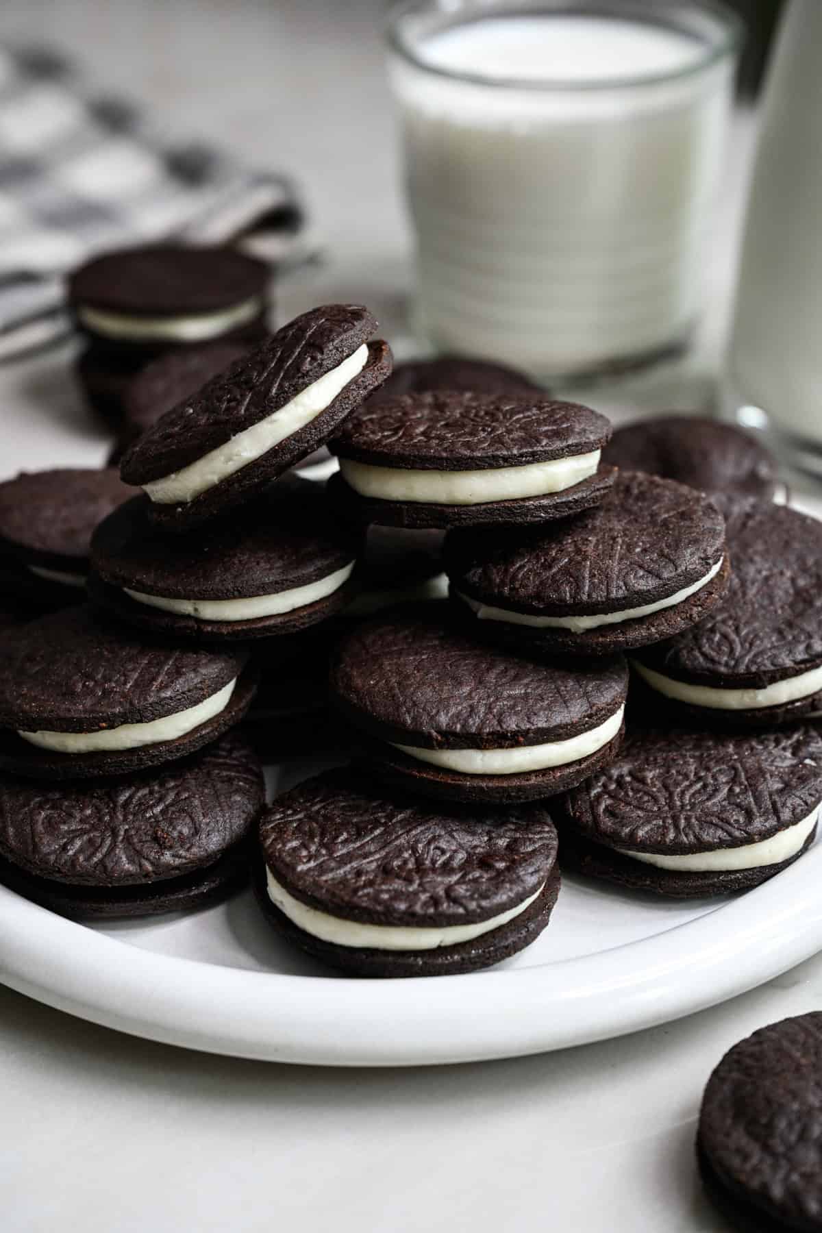 Close up side view of homemade oreos stacked on a white plate with a glass of milk behind. 