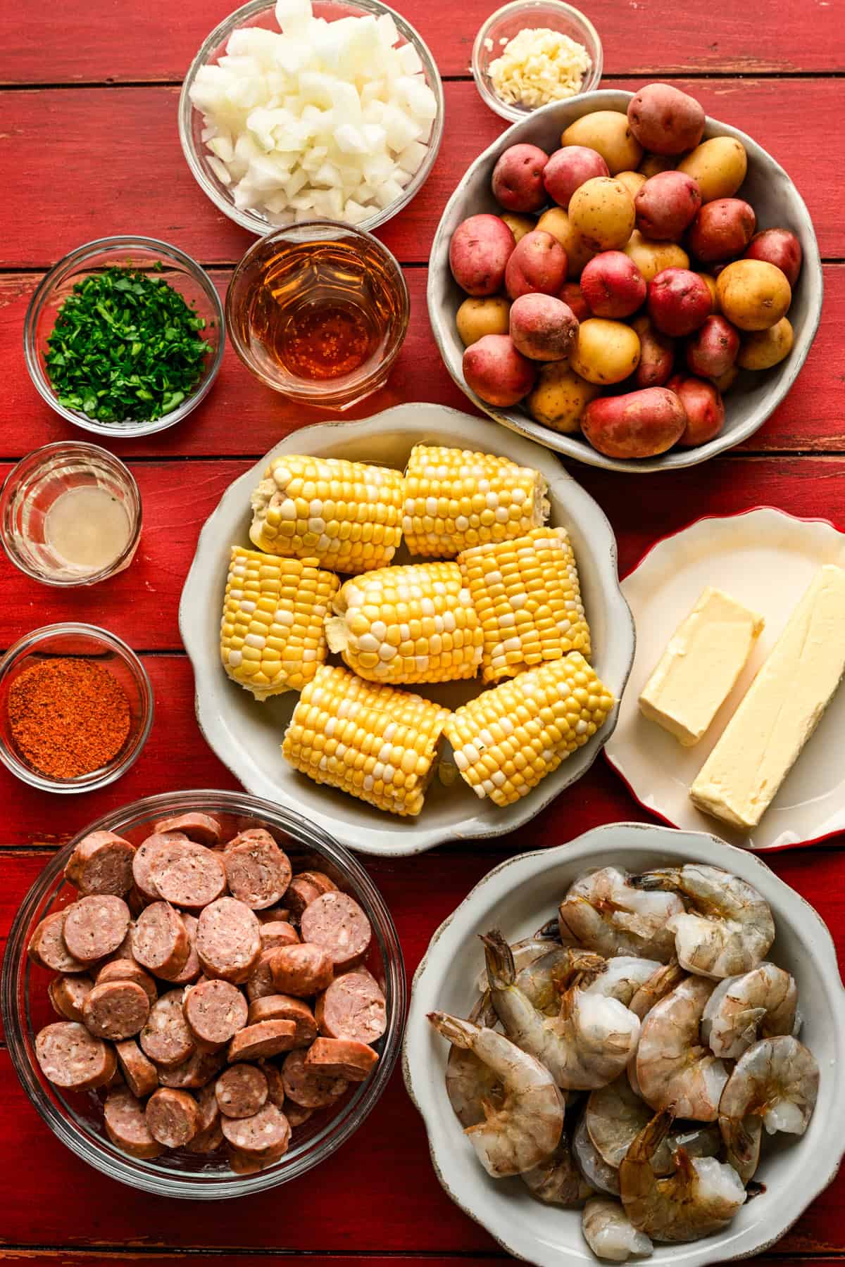 Overhead view of ingredients for sheet pan shrimp boil on a red table in separate bowls.