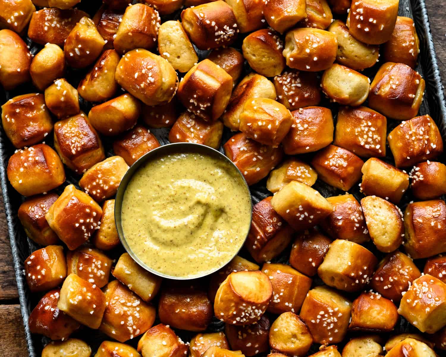Overhead view of a sheet pan piled with golden-brown soft pretzel bites topped with pretzel salt, with a small metal bowl of creamy mustard dip in the center.