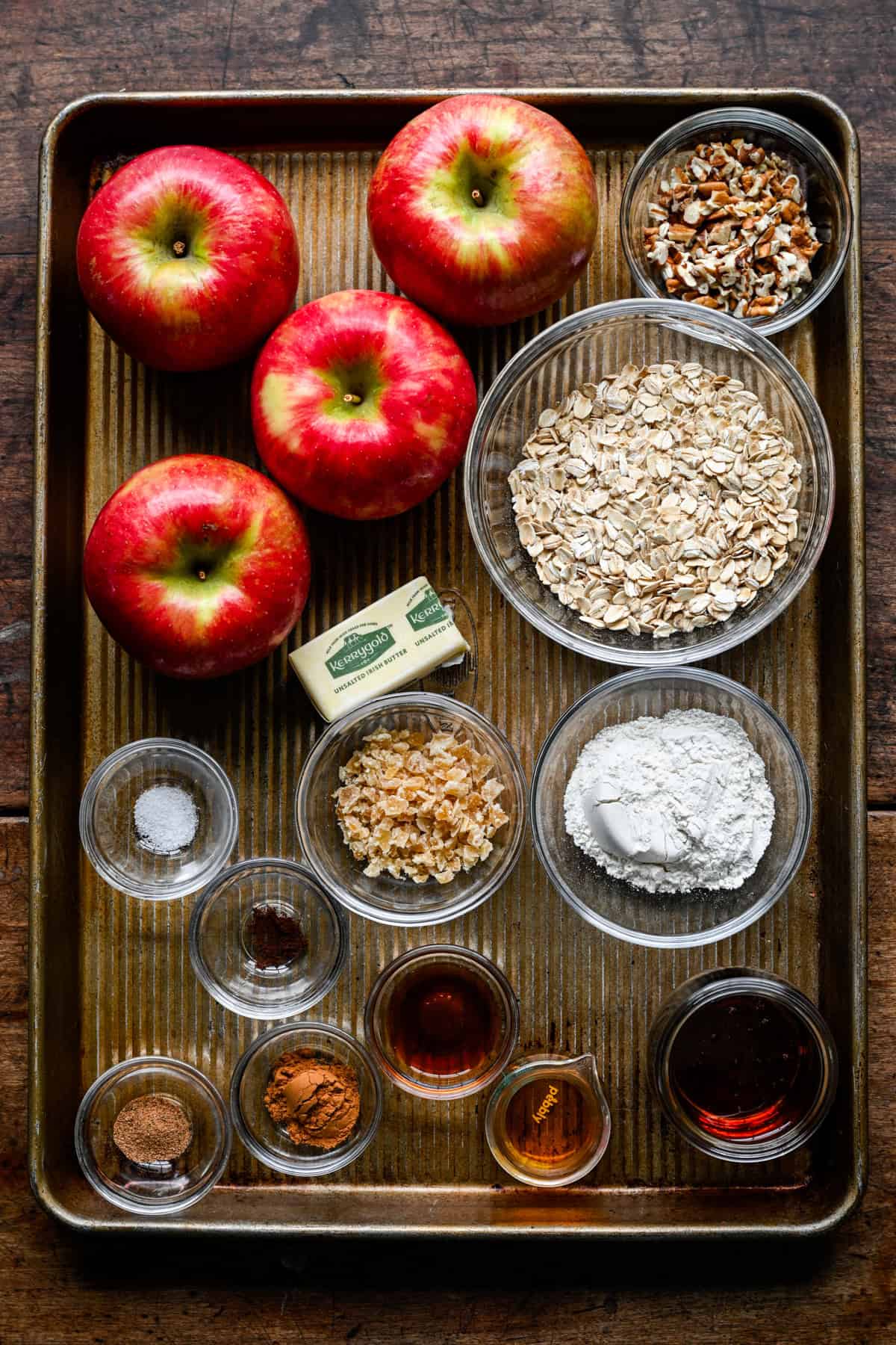 Ingredients for baked apples on a sheet pan: four honeycrisp apples, bowls of rolled oats, chopped pecans, flour, crystallized ginger, a stick of butter, ground spices, maple syrup and vanilla.