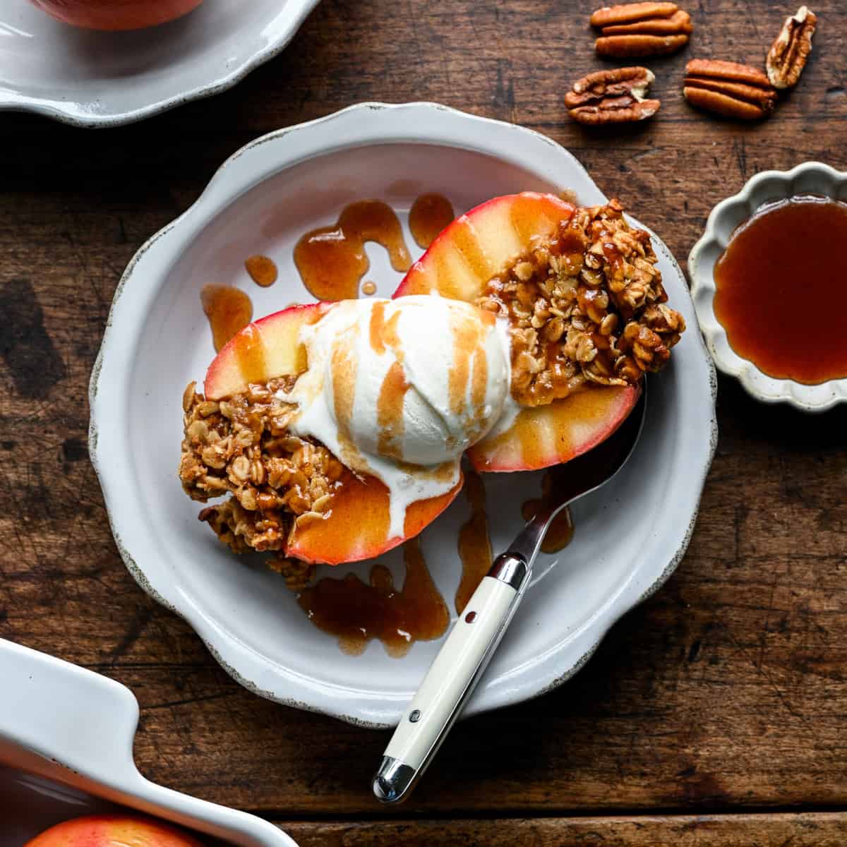 A baked apple cut in half on a plate with a scoop of vanilla ice cream, drizzled with date caramel, pecans and a small dish of the caramel alongside.