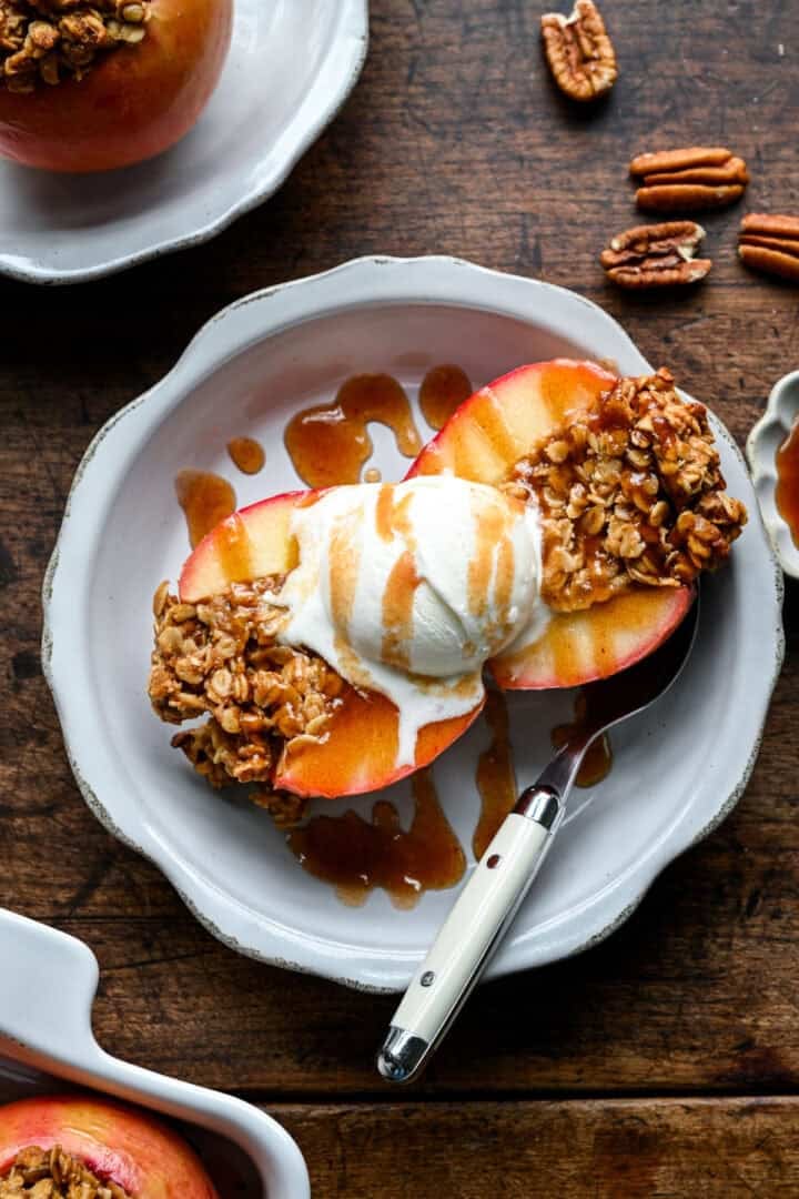 A halved baked apple with vanilla ice cream and date caramel, a second serving and scattered pecans in the background.