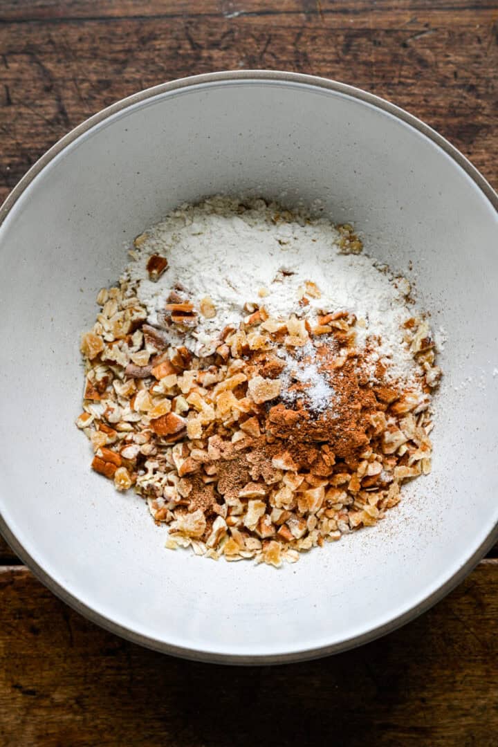 Rolled oats, chopped pecans, crystallized ginger, flour, cinnamon, nutmeg, cloves and salt in a white mixing bowl before stirring.