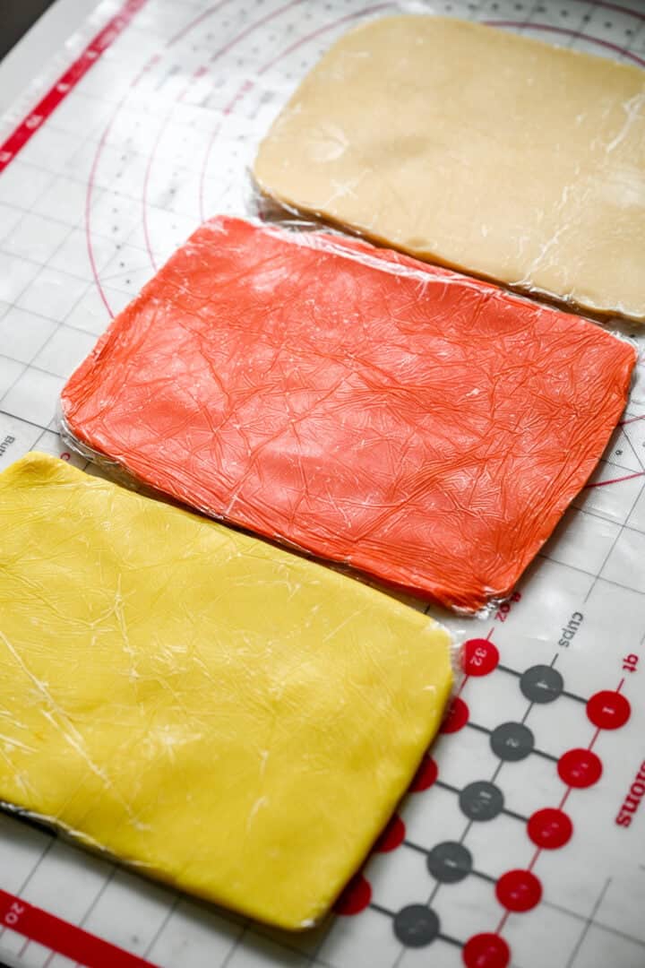 Three rolled-out rectangles of dough wrapped in plastic: yellow, orange and plain.