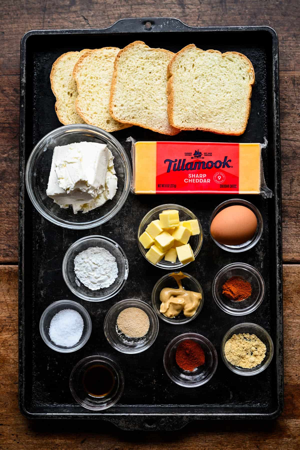 Overhead view of cheese dreams ingredients on a dark sheet pan: bread, cream cheese, a block of Tillamook sharp cheddar, cubed butter, an egg, mustard and small bowls of spices.