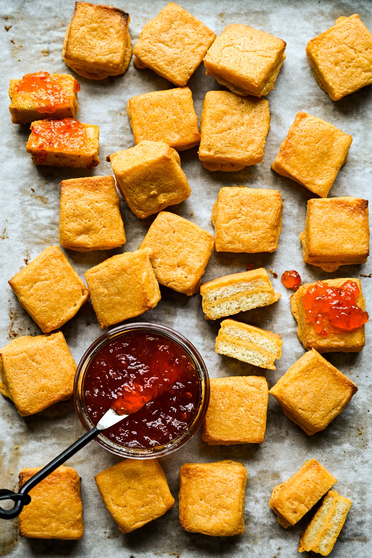 Overhead view of cheese dreams scattered on parchment around a jar of red pepper jelly with a spreader.