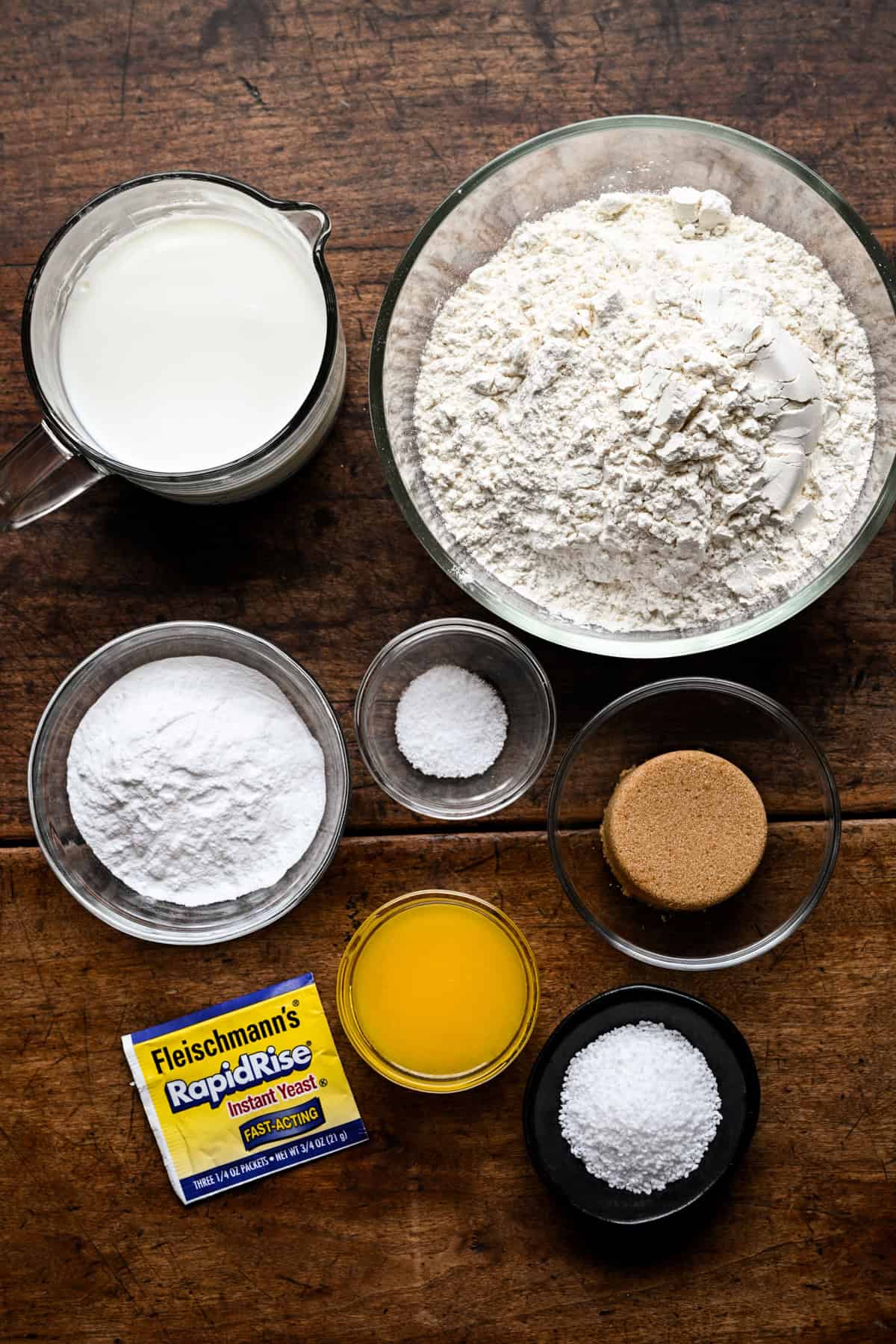 Ingredients for pretzel bites on a wooden table: a measuring cup of milk, a bowl of flour, small bowls of baking soda, salt, brown sugar and pretzel salt, melted butter, and a packet of instant yeast.