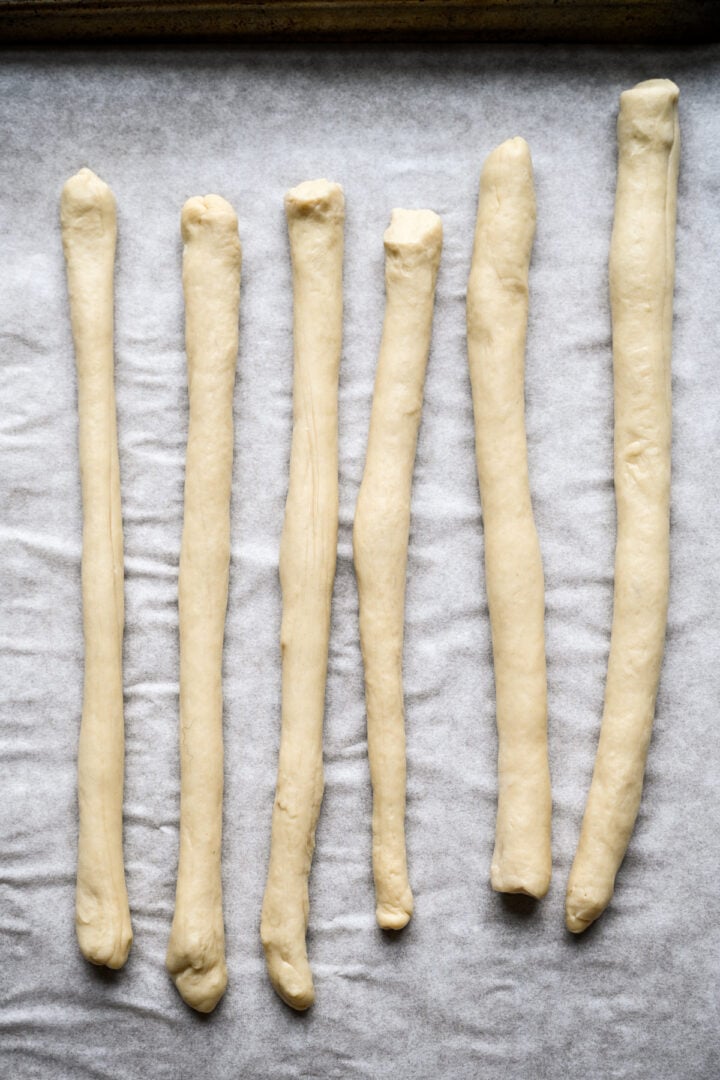 Six long ropes of pretzel dough rolled out side by side on parchment paper.