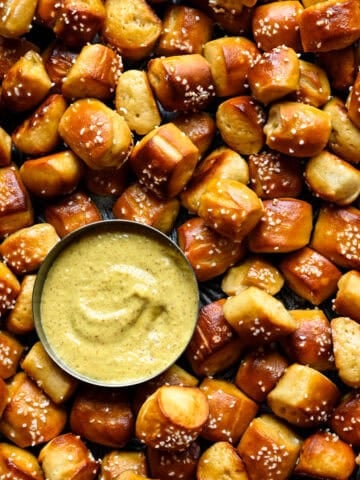 Close overhead view of salted soft pretzel bites heaped around a small metal bowl of creamy mustard dip with a swirl on top.