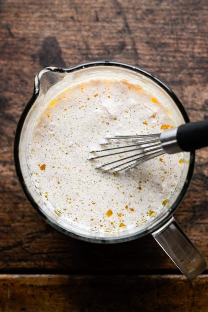 Melted butter being whisked into the foamy yeast mixture in a glass measuring cup.
