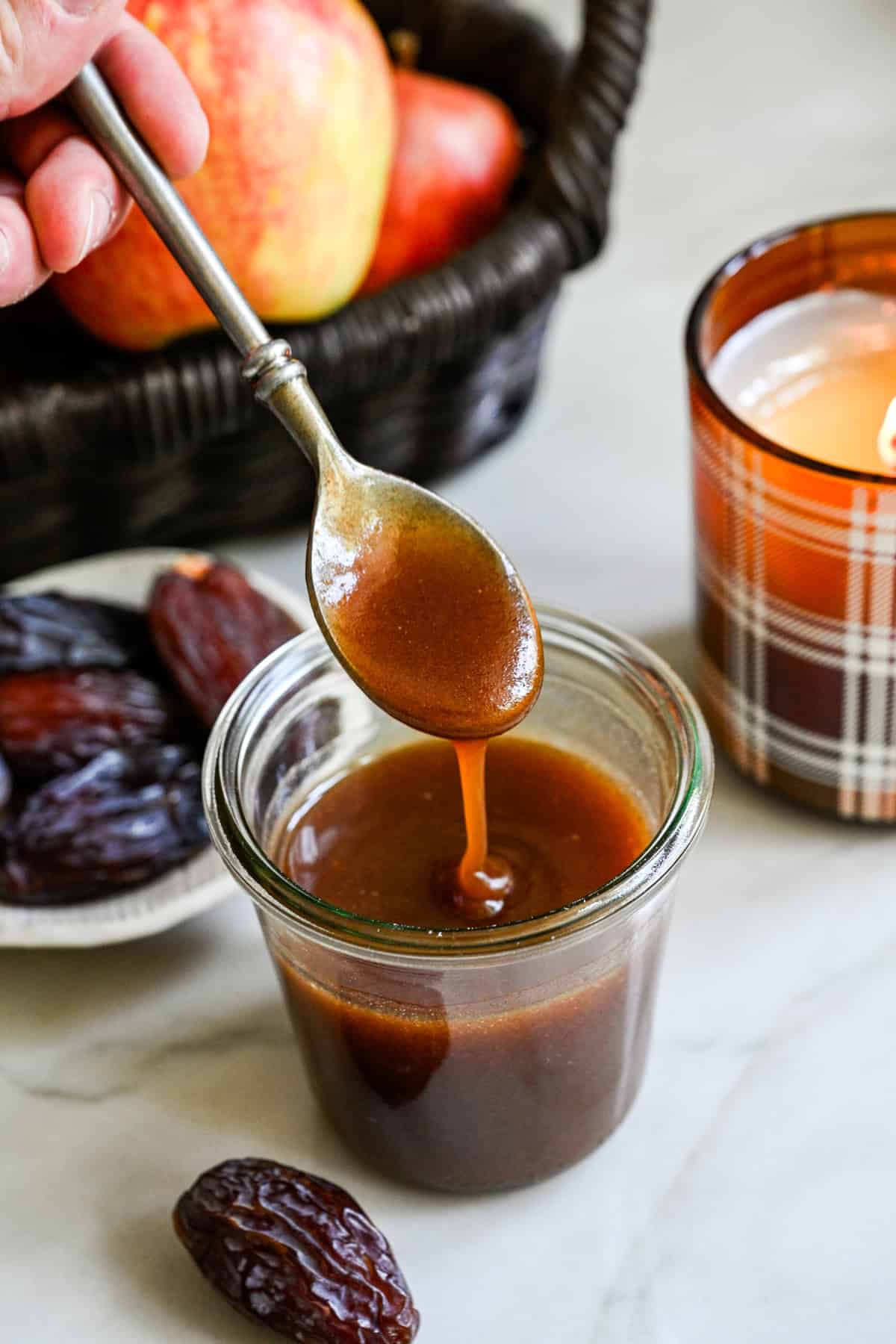 Close up view of date caramel sauce in a glass jar with a spoon lifted above that's drizzling caramel back into the jar. 