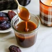 Close up view of date caramel sauce in a glass jar with a spoon lifted above that's drizzling caramel back into the jar.