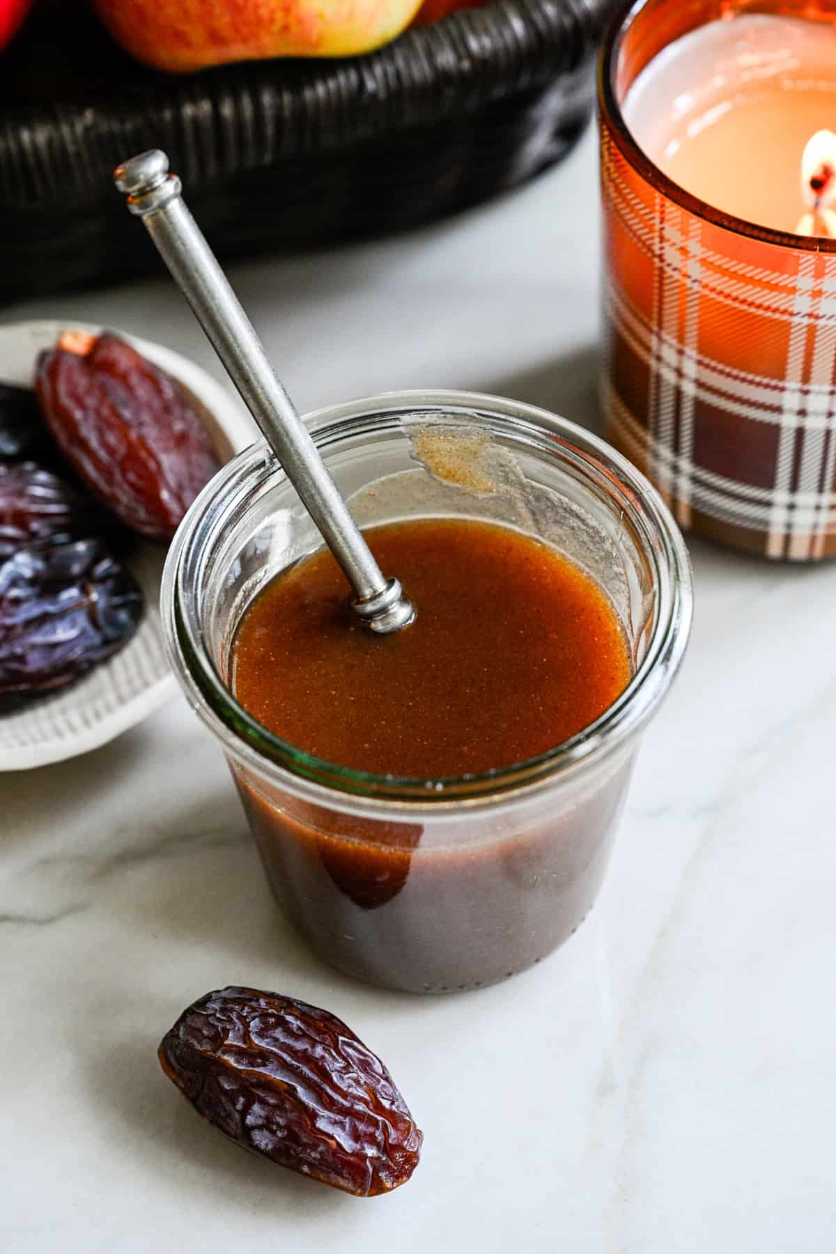 Date caramel sauce in a glass jar with a fall candle and a bowl of dates in the background.