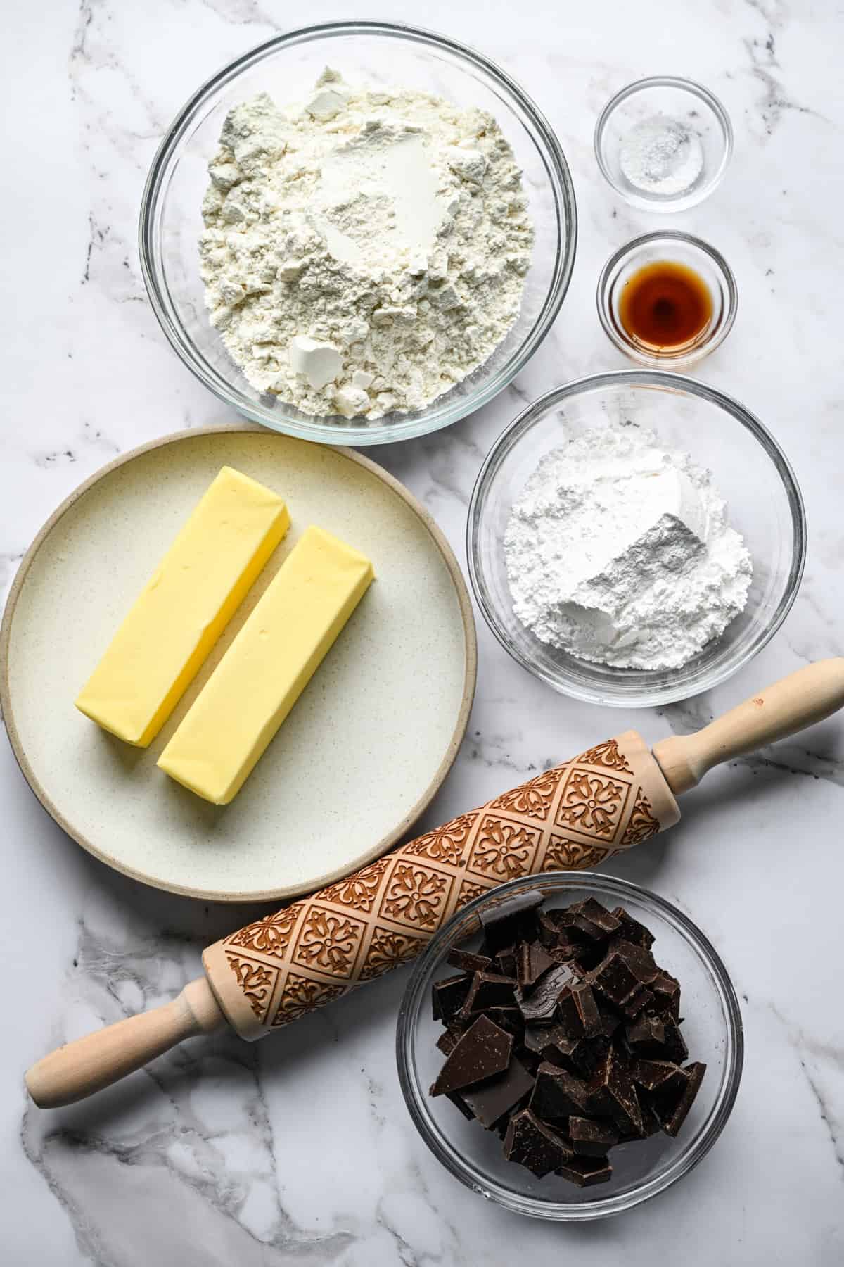 Glass bowls of flour, powdered sugar, chopped dark chocolate, vanilla, and salt on a marble counter with two sticks of butter on a plate and an embossed wooden rolling pin.