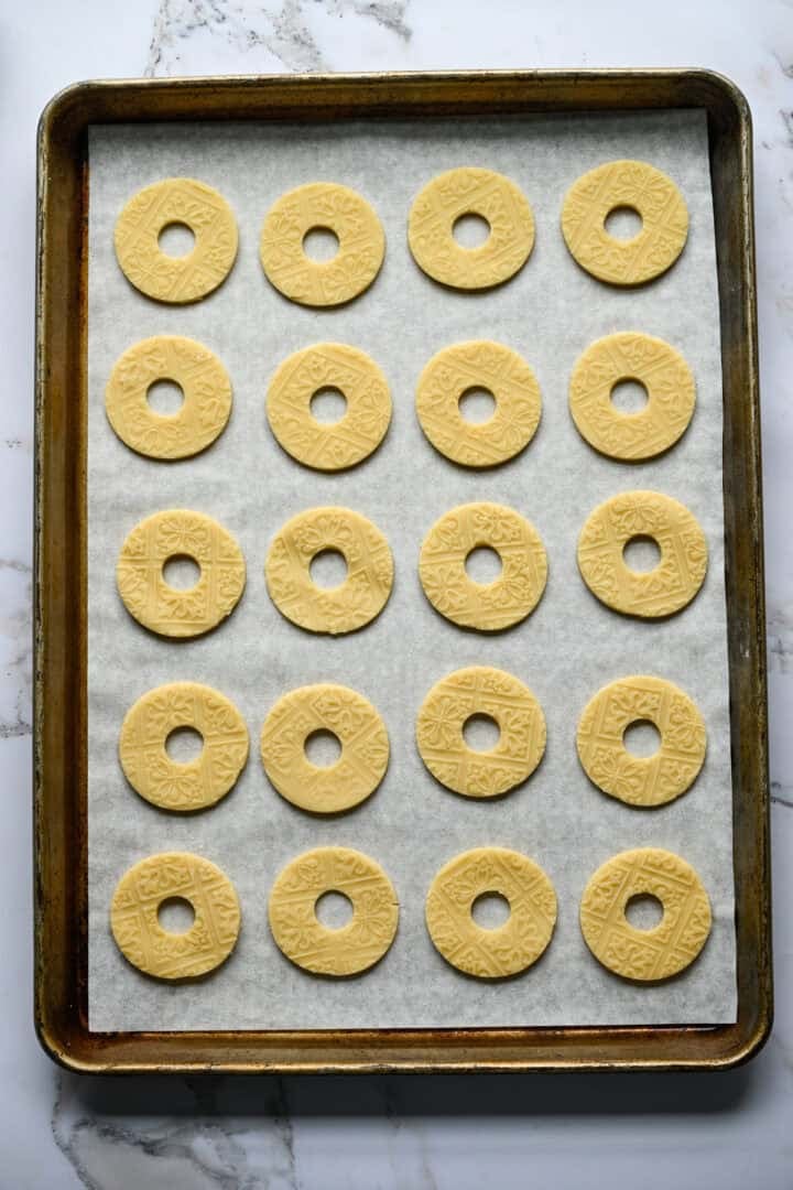 Twenty unbaked ring-shaped shortbread cookies with an embossed pattern lined up in rows on a parchment-lined baking sheet.