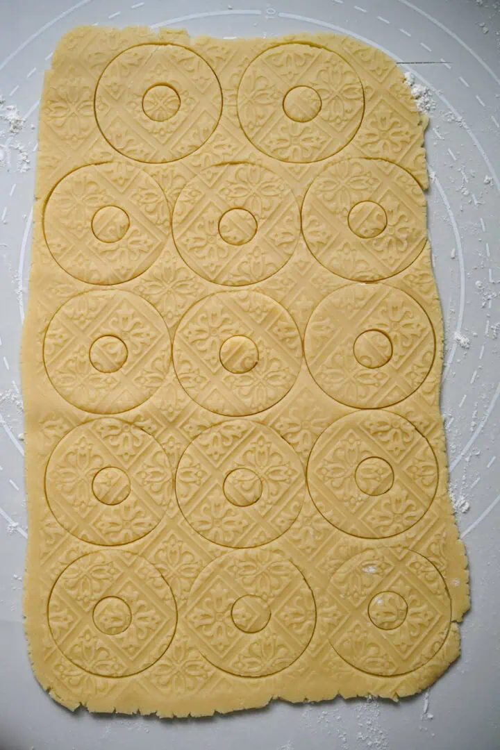 Rolled-out shortbread dough embossed with a floral tile pattern and cut into rounds with small center holes, before the cookies are lifted out.