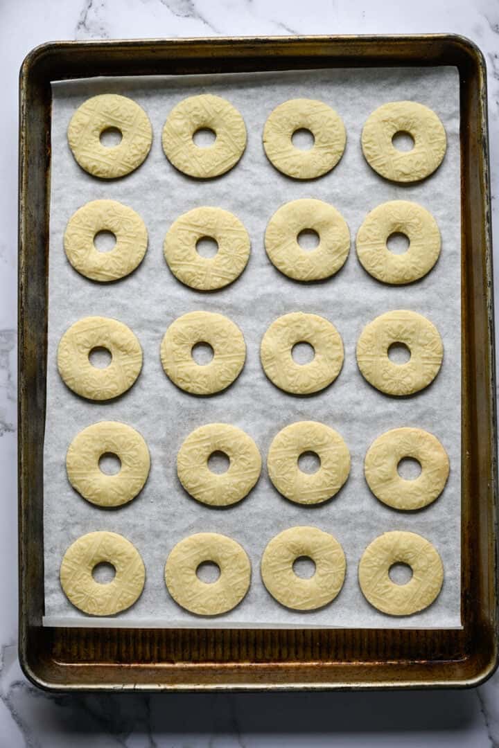 Twenty baked ring-shaped shortbread cookies, pale golden with an embossed pattern, on a parchment-lined baking sheet.