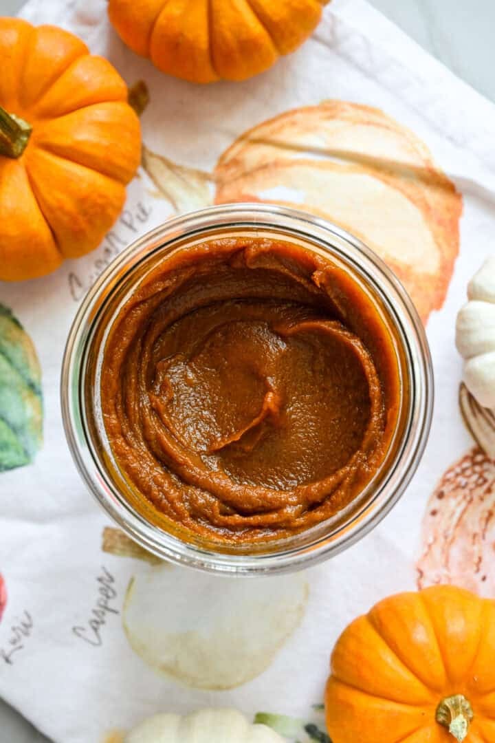 Close up view of a jar of homemade pumpkin butter on a fall-themed linen next to mini pumpkins for decor.