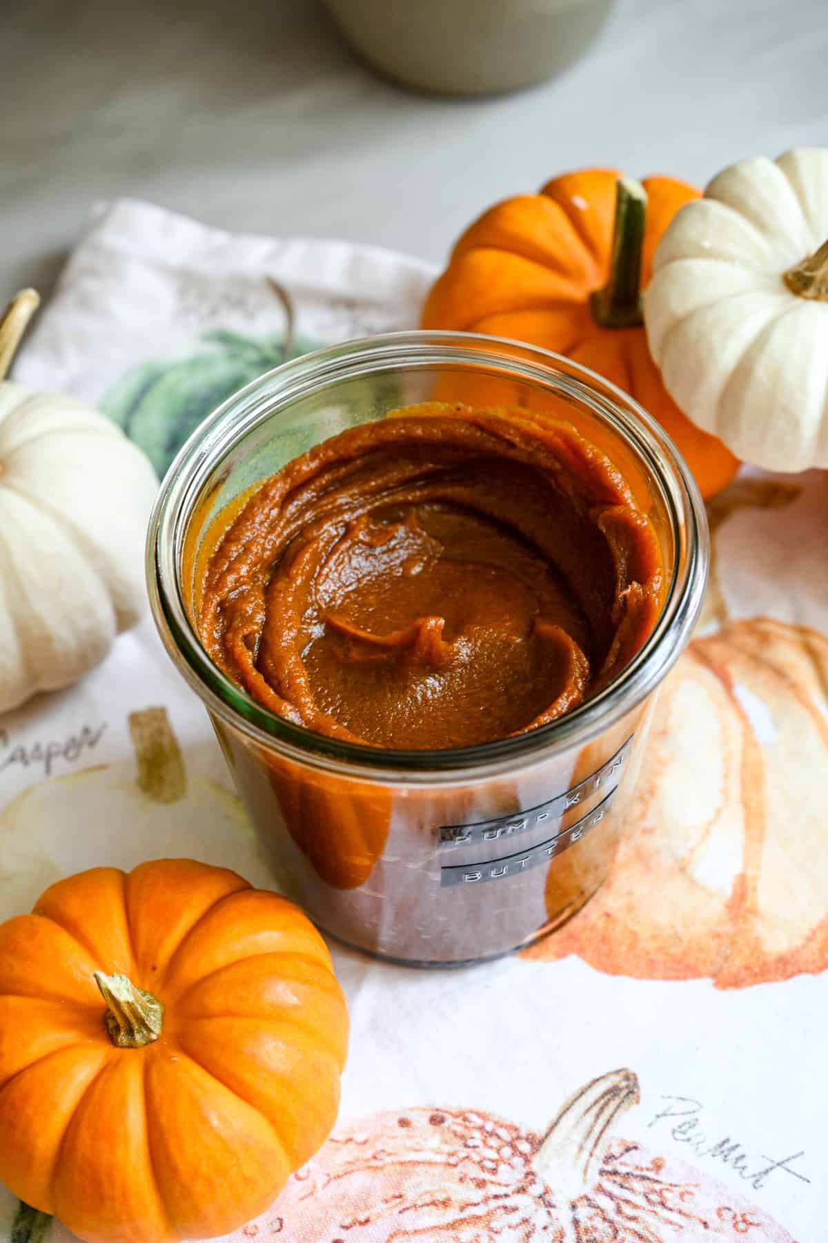 Close up view of a jar of homemade pumpkin butter on a fall-themed linen next to mini pumpkins for decor.