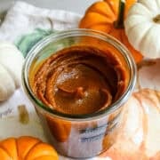 Close up view of a jar of homemade pumpkin butter on a fall-themed linen next to mini pumpkins for decor.