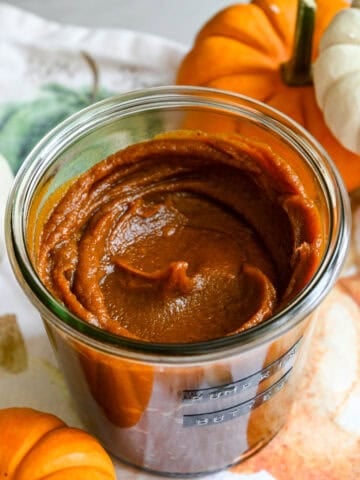 Close up view of a jar of homemade pumpkin butter on a fall-themed linen next to mini pumpkins for decor.