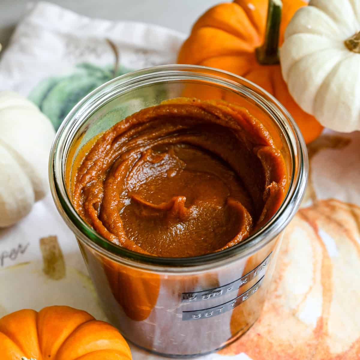Close up view of a jar of homemade pumpkin butter on a fall-themed linen next to mini pumpkins for decor.
