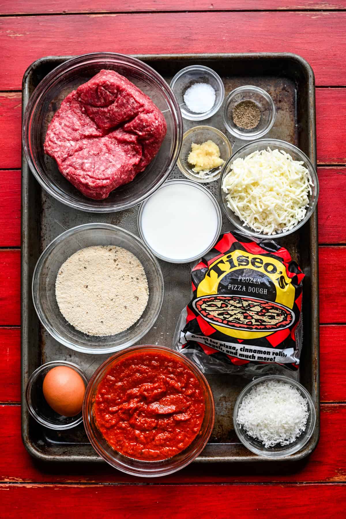 Overhead view of meatball sub bite ingredients on a sheet pan: ground beef, breadcrumbs, milk, egg, garlic, mozzarella, parmesan, marinara and Tiseo's frozen pizza dough.