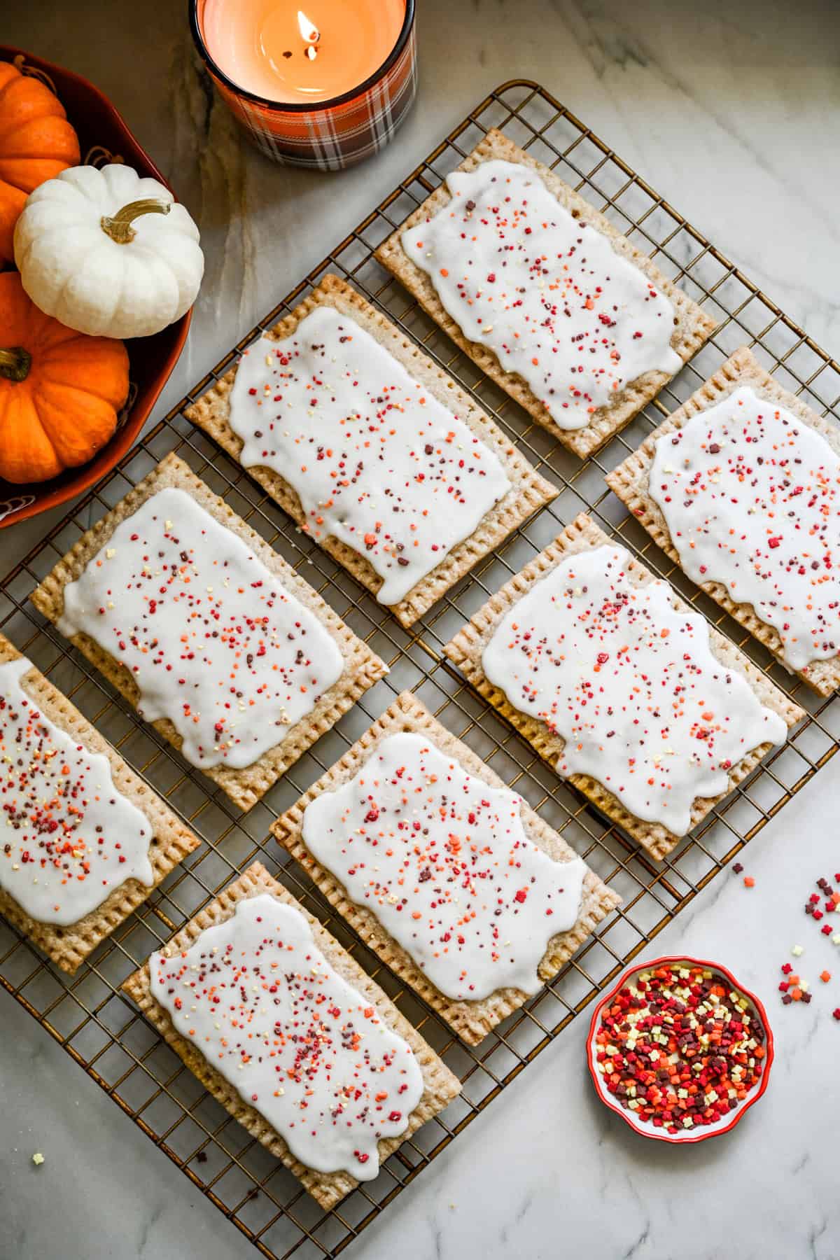 Overhead view of homemade pumpkin pie pop tarts on a wire rack with mini pumpkins and a fall candle on the side. 