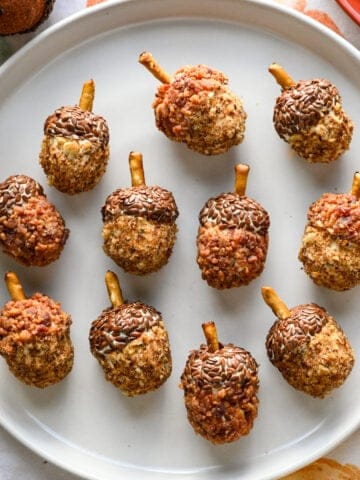 Overhead view of eleven acorn cheese balls coated in flax seeds, bacon and pecans arranged on a white plate.