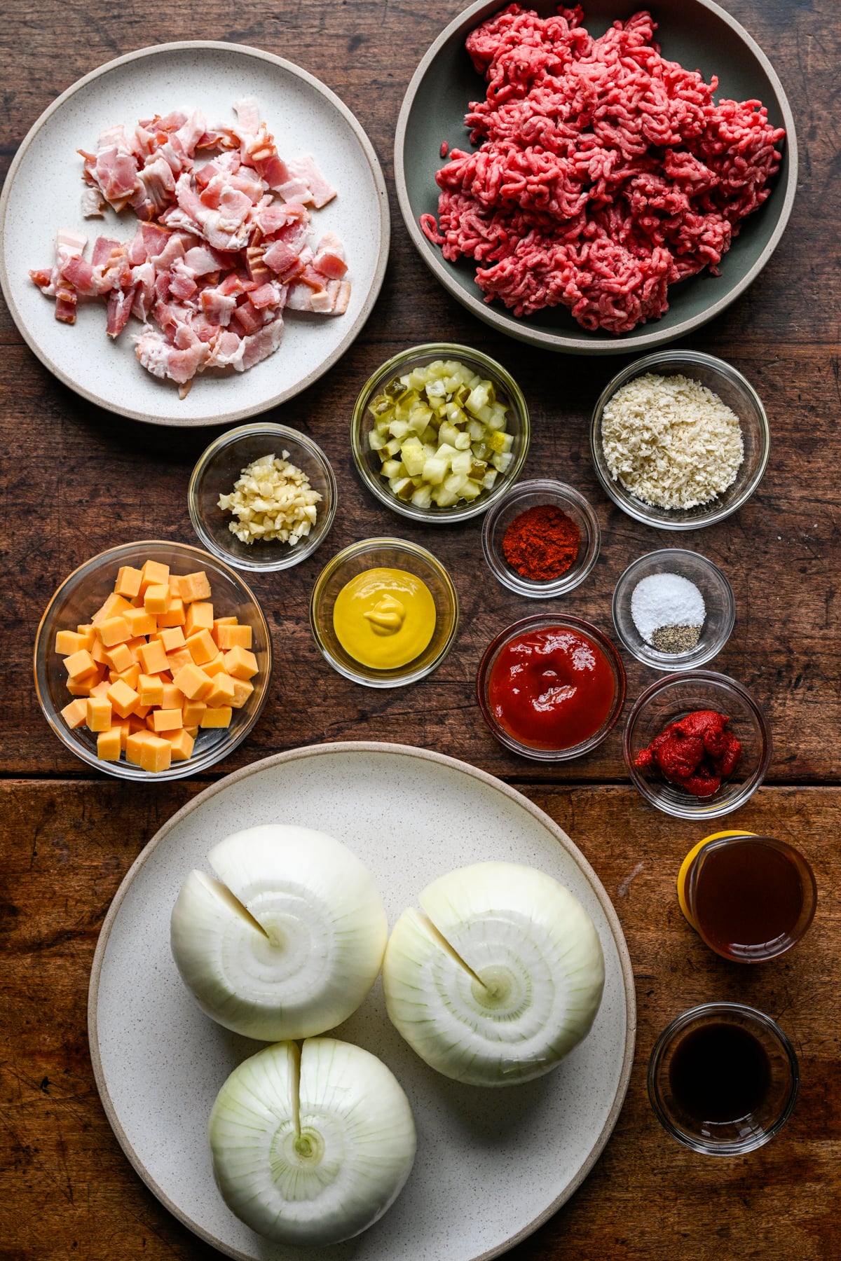 Overhead view of ingredients for bacon cheeseburger stuffed onions on a wood table: three peeled whole onions, raw ground beef, chopped bacon, cubed cheddar, chopped pickles, breadcrumbs, garlic, mustard, ketchup, tomato paste, smoked paprika, salt and pepper, beef broth and Worcestershire sauce