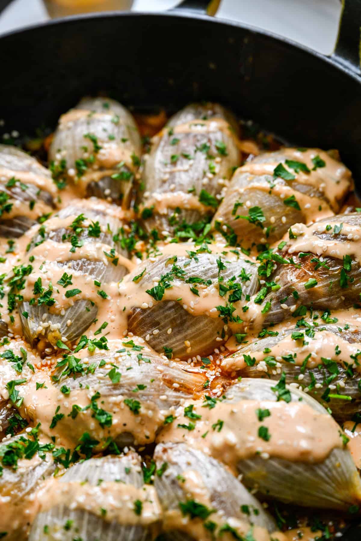 Close up view of baked bacon cheeseburger stuffed onions in a cast iron skillet drizzled with Thousand Island dressing, sesame seeds and parsley