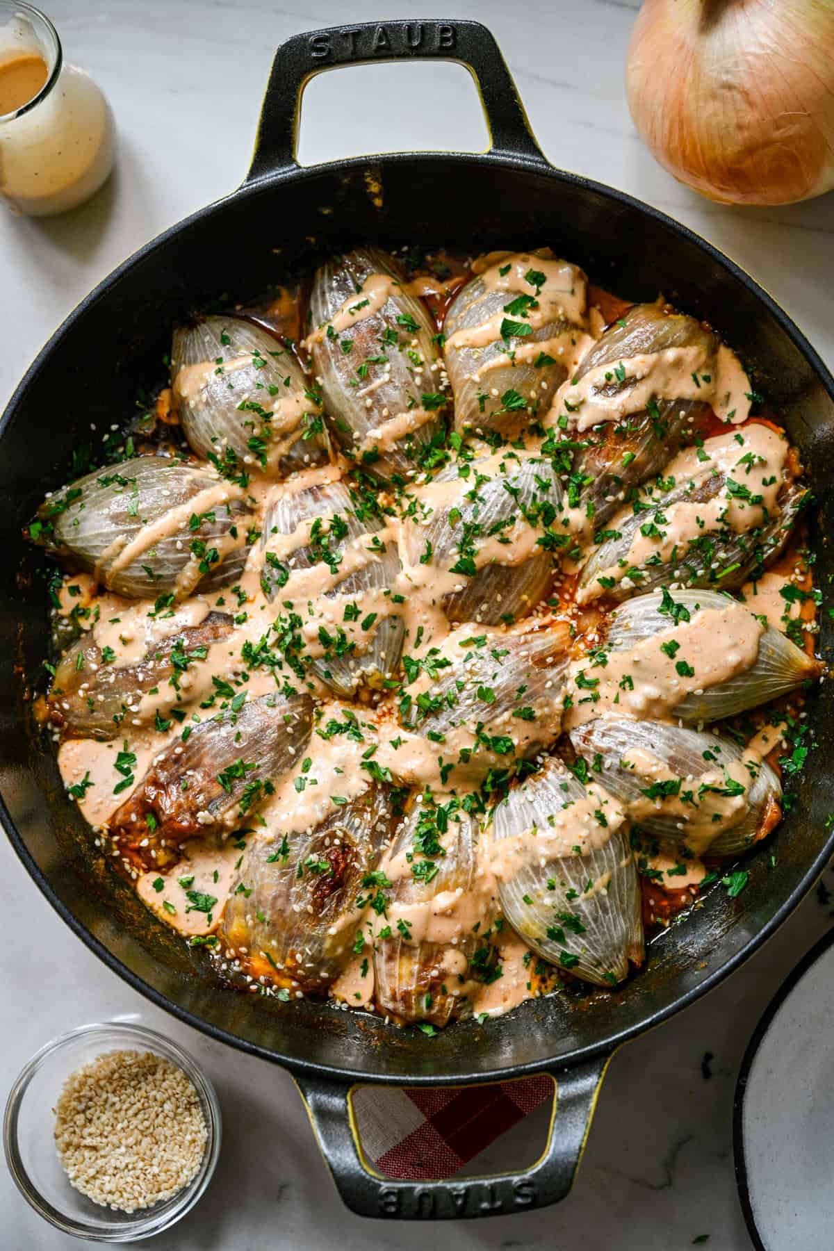 Overhead view of bacon cheeseburger stuffed onions in a Staub cast iron skillet topped with Thousand Island dressing, parsley and sesame seeds, with a jar of dressing and a bowl of sesame seeds beside it