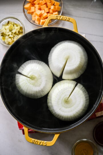 Overhead view of three whole peeled onions, each with a slit cut to the center, simmering in a yellow Staub pot