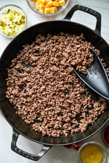 Overhead view of browned ground beef in a Staub cast iron skillet with a black spatula, bowls of pickles and cubed cheddar nearby