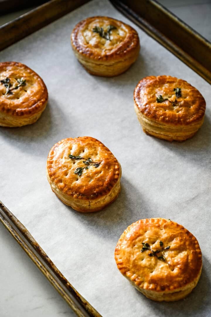 45 degree view of five baked golden brown chicken and leek hand pies on a parchment-lined sheet pan, each topped with a thyme sprig.