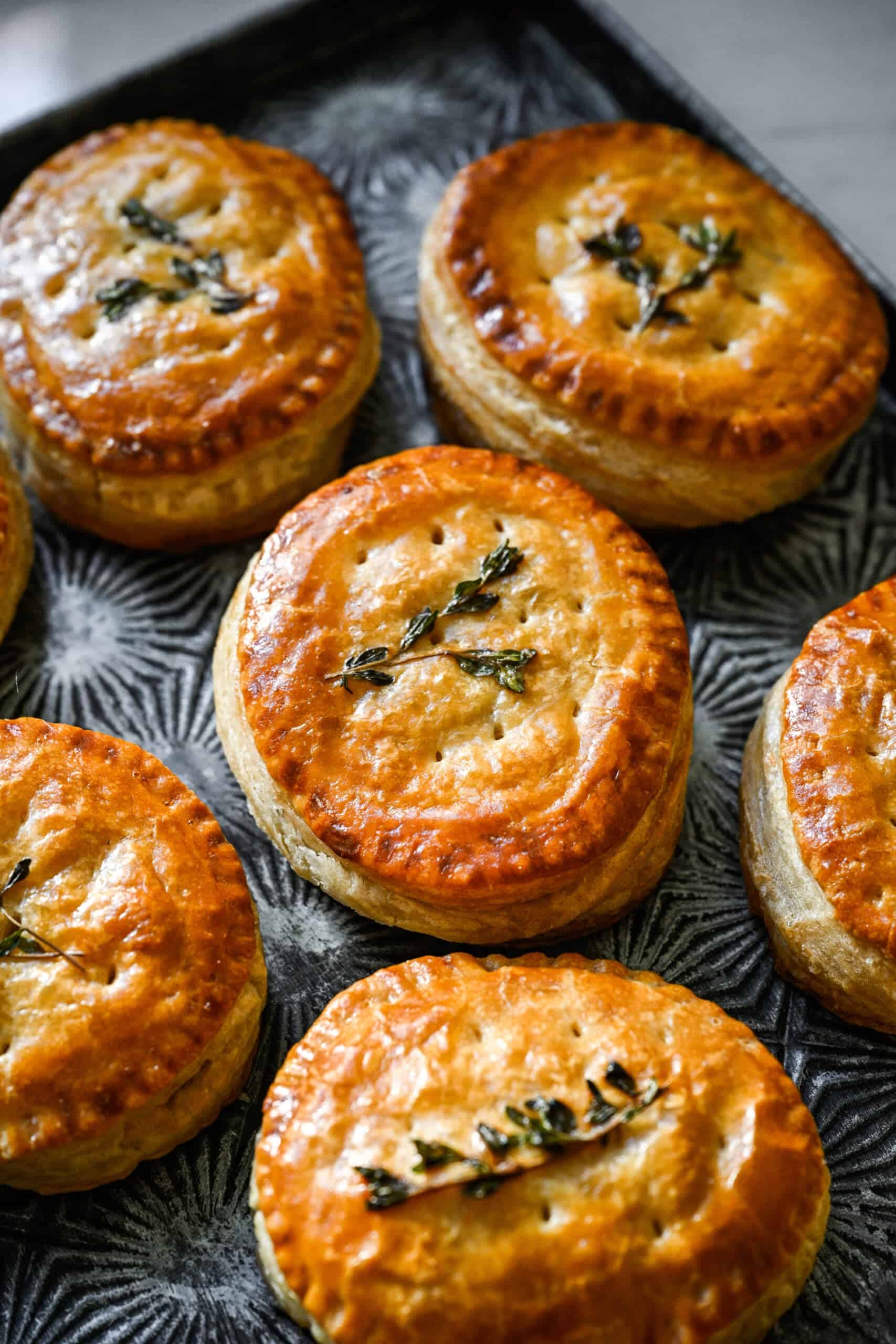 Close up view of baked golden puff pastry hand pies with thyme on top, arranged on a dark patterned metal tray.