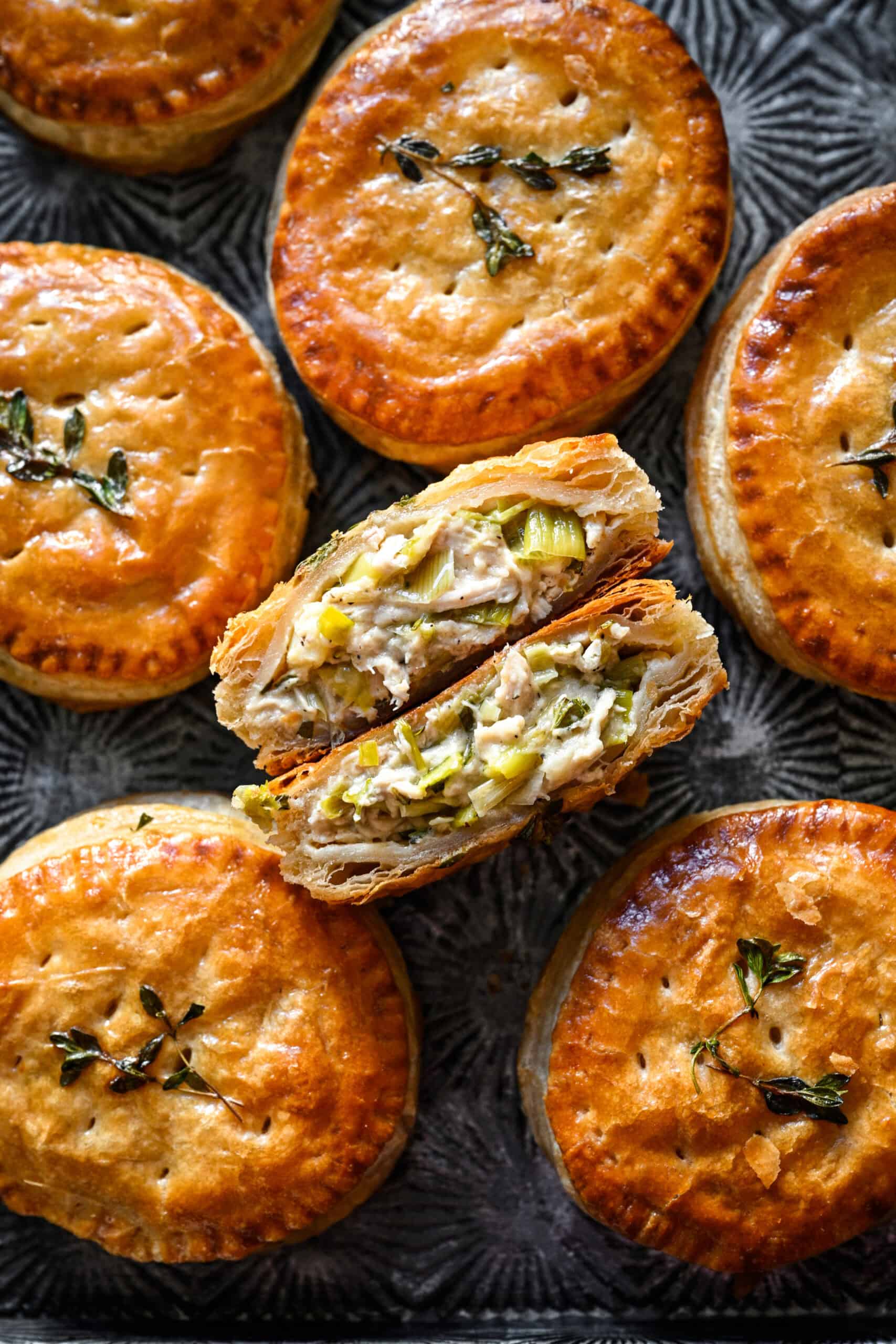 Overhead view of a chicken and leek hand pie cut in half to show the creamy chicken and leek filling and flaky layers, surrounded by whole hand pies on a dark tray.