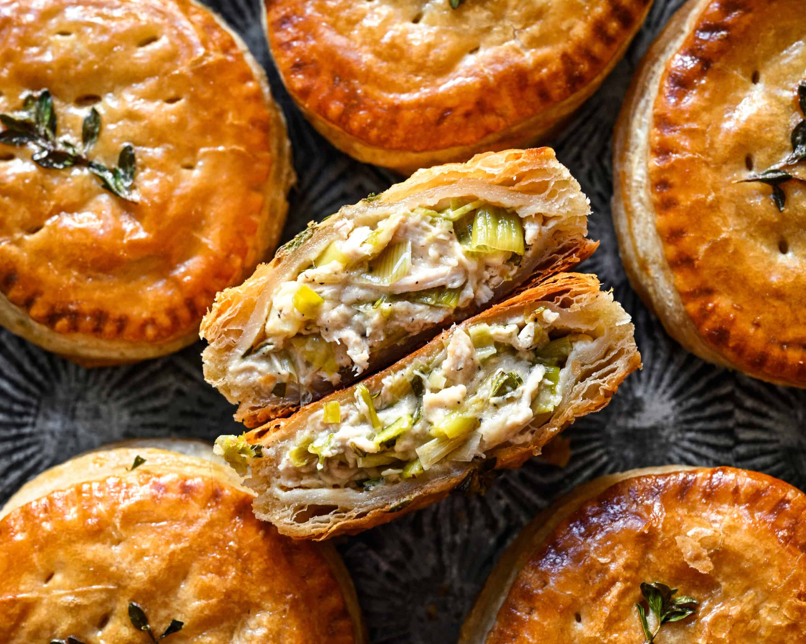 Close up view of the two halves of a chicken and leek hand pie stacked to show the creamy filling and flaky puff pastry layers, with whole pies around it.