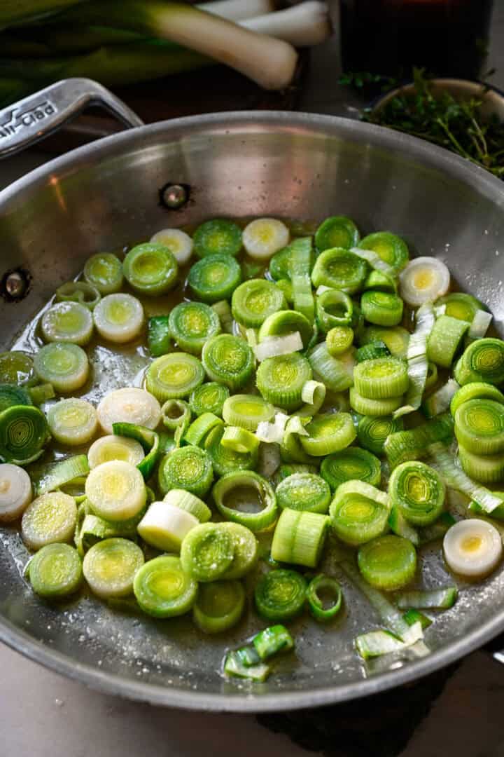 45 degree view of sliced leek rounds starting to cook in melted butter in a stainless steel skillet, with whole leeks and a bowl of thyme behind.