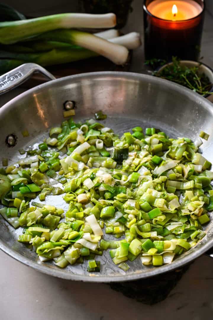 45 degree view of softened, wilted chopped leeks in a stainless steel skillet, with whole leeks and a lit candle behind.