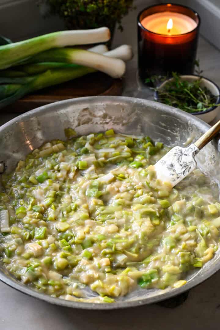 45 degree view of cooked leeks coated in a thick, gravy-like sauce in a stainless steel skillet, being stirred with a floral spatula.
