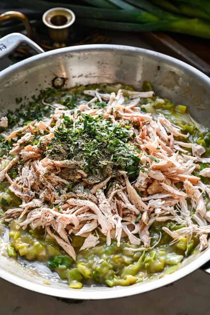 45 degree view of shredded chicken and chopped fresh herbs piled on top of the creamy leek mixture in a skillet before stirring.