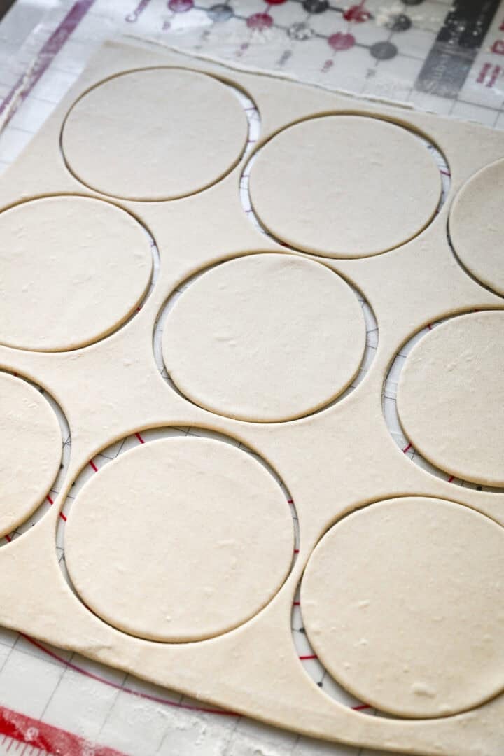 45 degree view of a sheet of rolled puff pastry on a gridded pastry mat with nine round circles cut out of it.
