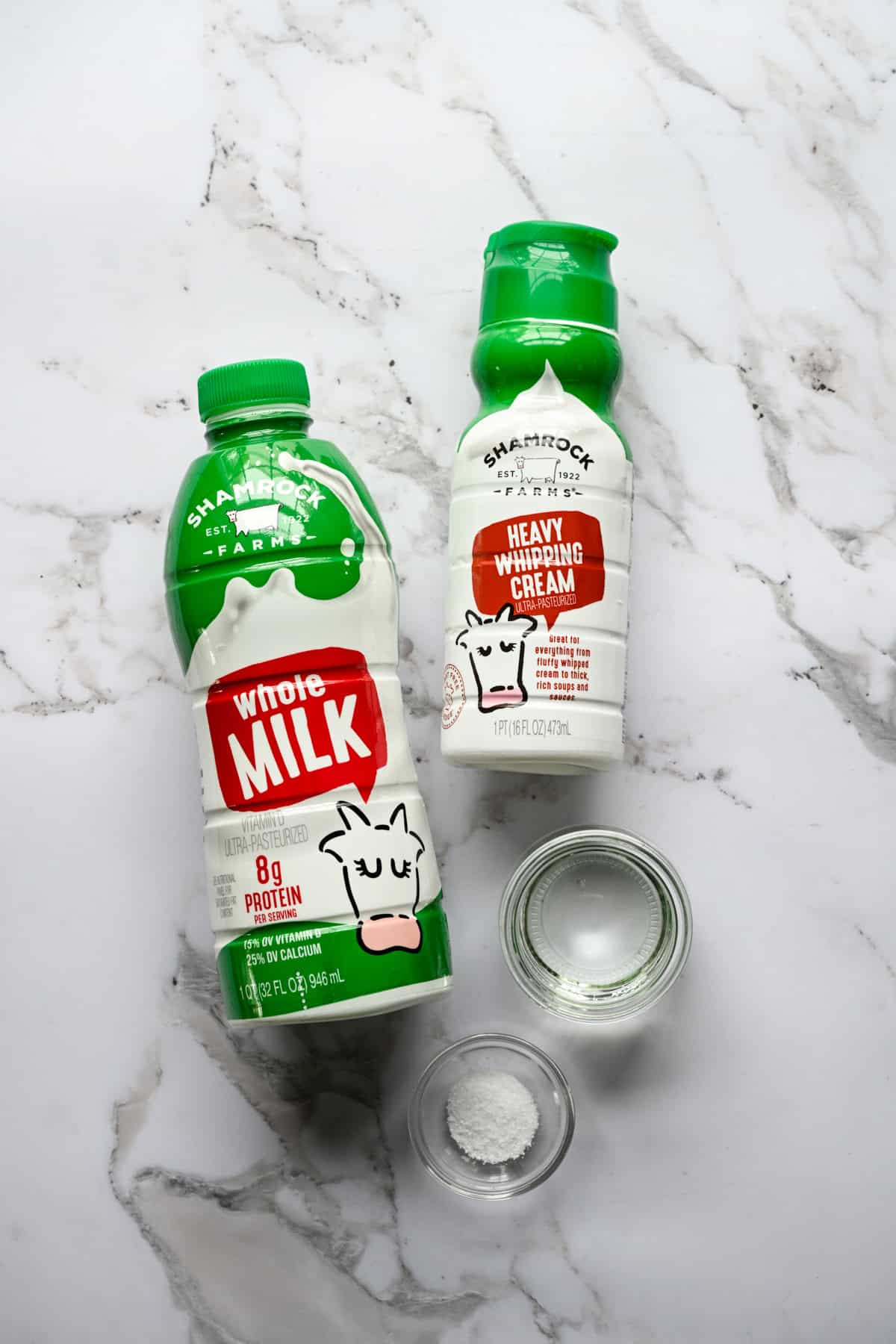 Overhead view of ingredients for homemade cottage cheese: a bottle of Shamrock Farms whole milk, Shamrock Farms heavy whipping cream, and small glass bowls of white vinegar and salt on marble